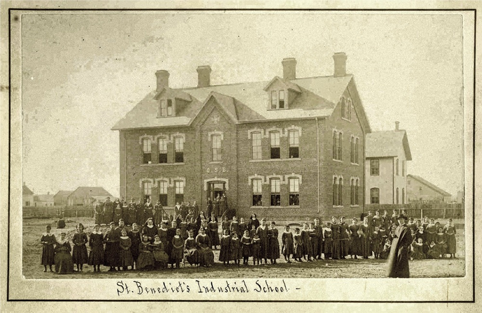 A priest and students posing in front of Saint Benedict's Indian Industrial School, newly reconstructed in 1886.